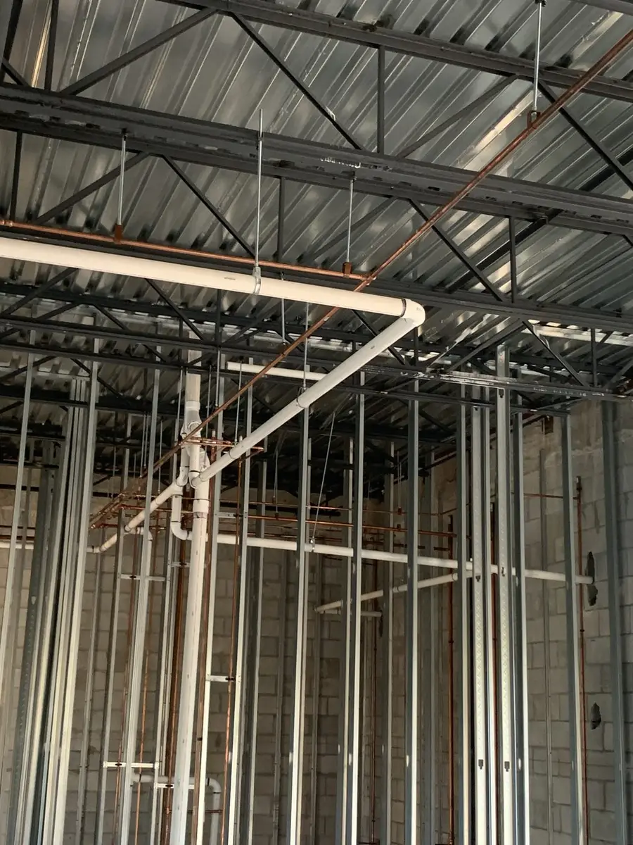 Overhead PVC drain and vent lines run through steel roof framing on a commercial shell
